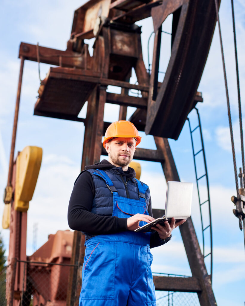 Vertical portrait of oil man, wearing blue overalls and orange helmet, with a laptop, standing with his back to an oil rig, checking oil pumping unit, making notes in his computer