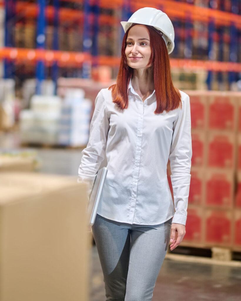 Warehouse. Confident smiling young woman in white blouse and safety helmet with laptop standing looking to side at large lighted warehouse